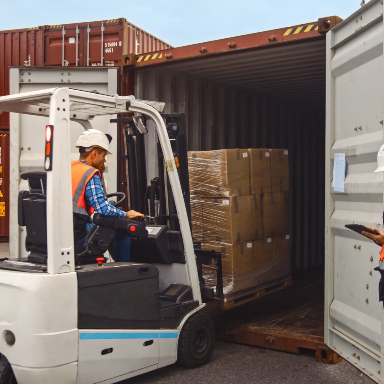 Forklift Driver Loading a Shipping Cargo Container with a Full Pallet with Boxes in Logistics Port Terminal. Latin Female Industrial Supervisor and Safety Inspector with Tablet Managing the Process.