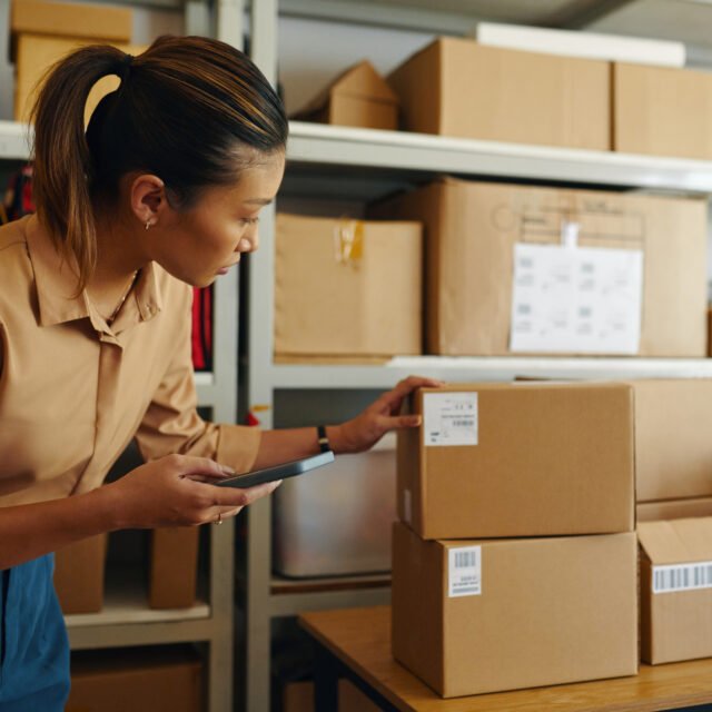 Young woman checking barcode on parcel with smartphone in storage room